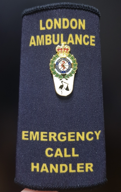 Epaulette of the London Ambulance Service, reading LONDON AMBULANCE at the top and EMERGENCY CALL HANDLER at the
  bottom. Pinned onto the epaulette is a badge showing the LAS logo, under which is a white inverted teardrop shape
  bearing a small embossed image of a stork.
